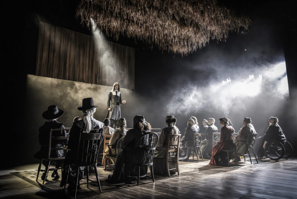 Performers sit on various wooden chairs facing a man dressed in pilgrim clothing.