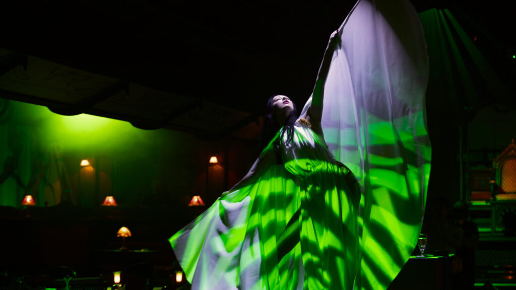 The Lost Estate – Chat Noir: a performer in an elegant dress wafts the long fabric as rich green gobo light washes over her