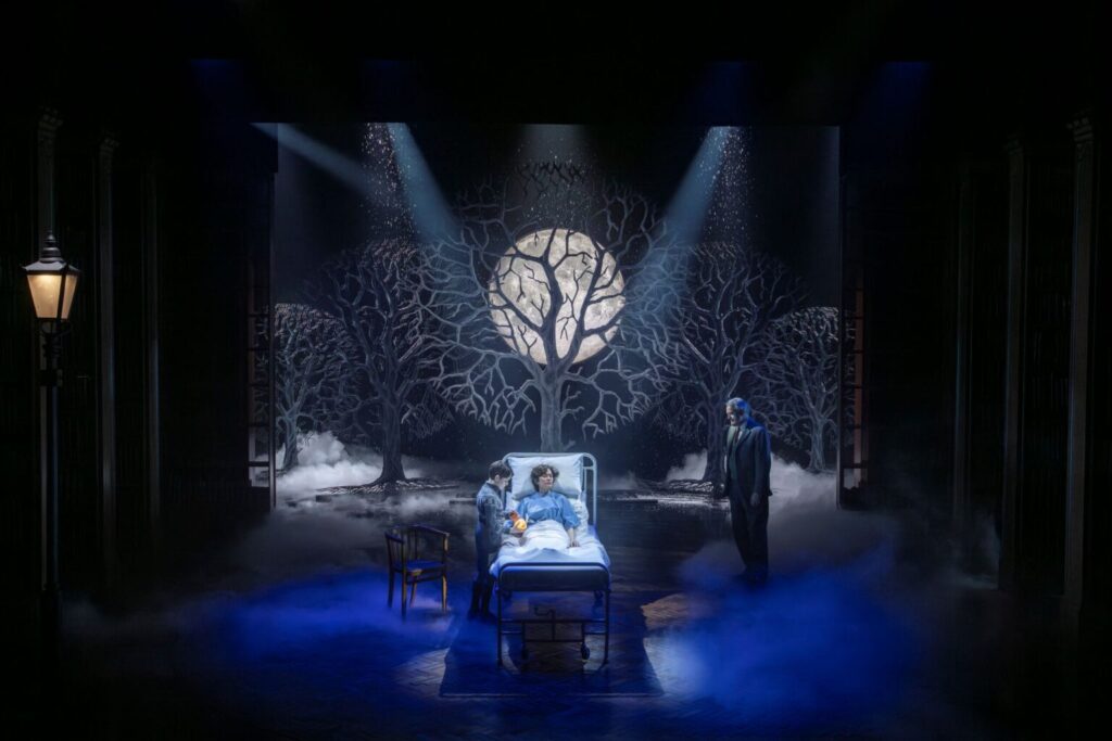 Scene from Shadowlands at the Aldwych Theatre: a young boy and an older man stand beside a bed on a fog covered stage, lit in blue tones with a moon glowing through silhouetted trees.