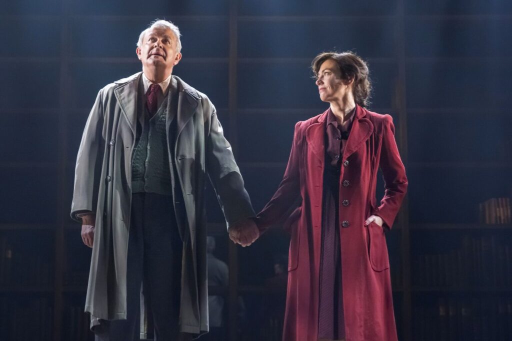 Scene from Shadowlands at the Aldwych Theatre: an older man and woman stand centre stage holding hands in long coats; he looks upward as she gazes at him lovingly.